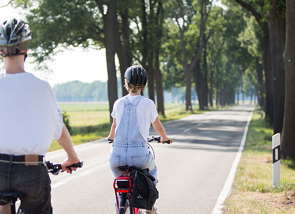 Zwei Personen mit Fahrradhelmen fahren auf einer schmalen Landstraße ohne separaten Radweg.